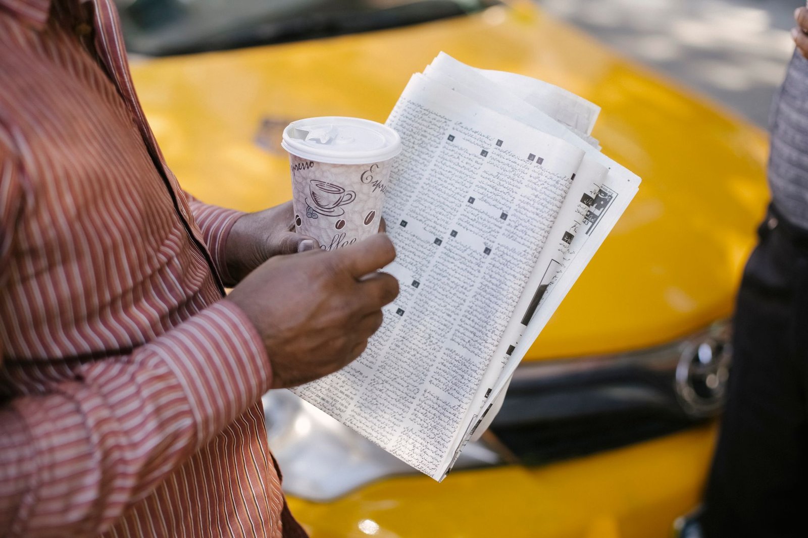 From above side view of crop anonymous ethnic male cab driver with coffee to go and newspaper talking to coworker on street —