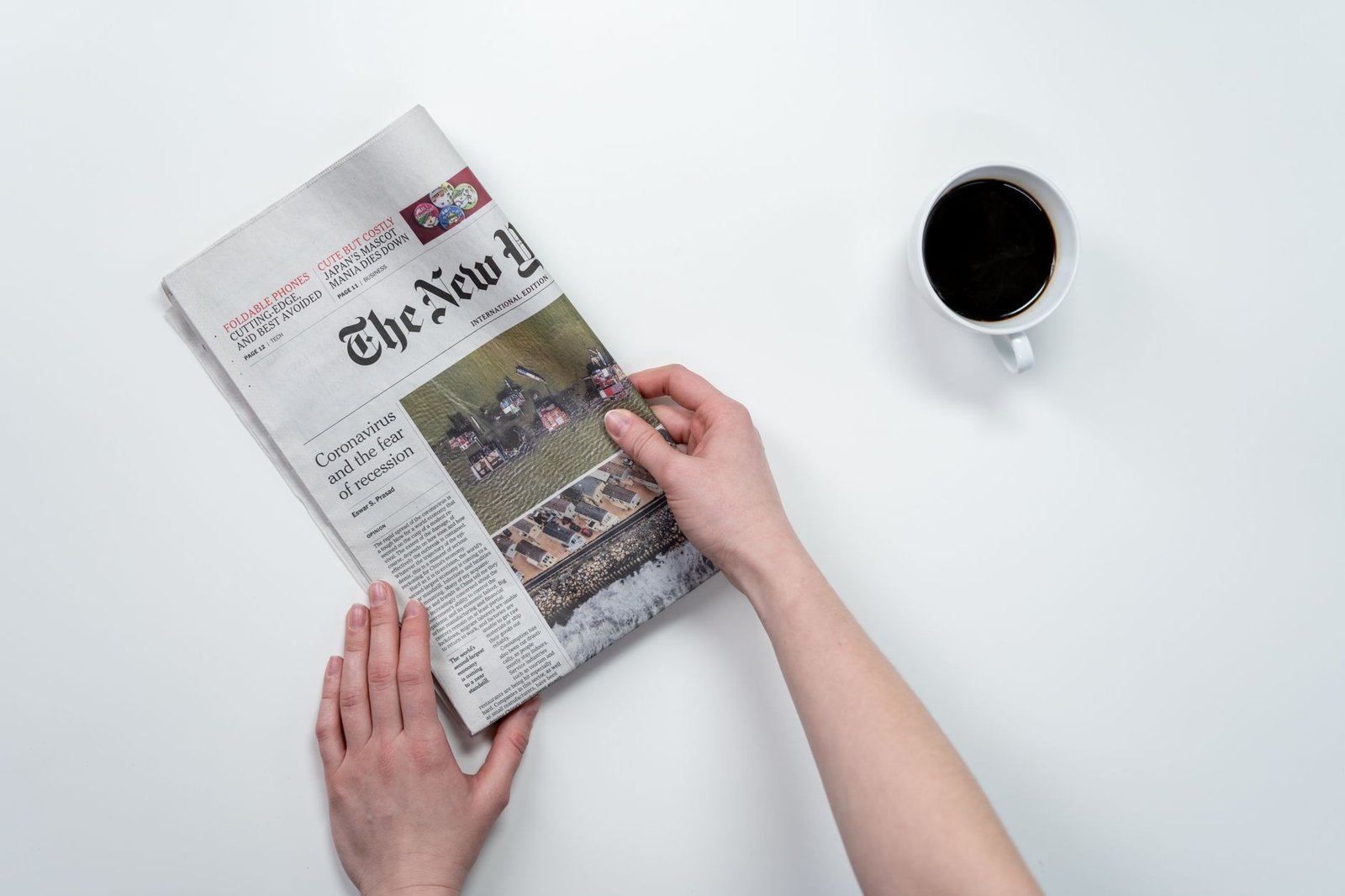 Flat lay of hands holding The New York Times newspaper with coffee on a white table. — Beyond Restrictions Experience Freedom