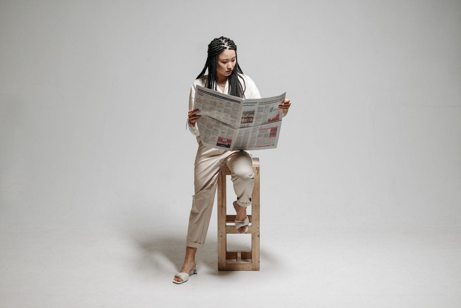A woman sits on a stool with a braided hairstyle, reading a newspaper in a minimalistic studio. — Examine Plans That have Dea